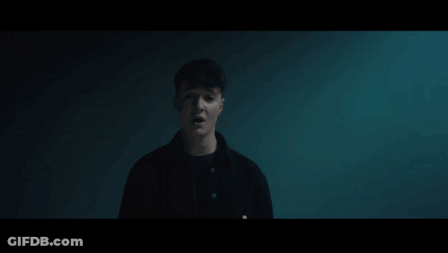 A Man In A Black Jacket Is Standing In A Dark Room Looking At The Camera GIF