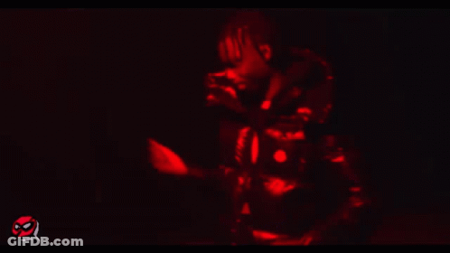 A Man In A Black Jacket Is Standing In A Dark Room With A Red Light Behind Him GIF