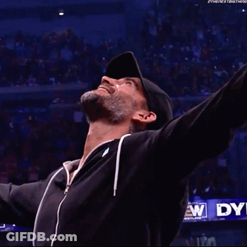 A Man In A Black Jacket Is Standing In A Wrestling Ring With His Arms Outstretched . GIF