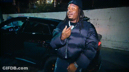 A Man In A Black Jacket Is Standing In Front Of A Car . GIF