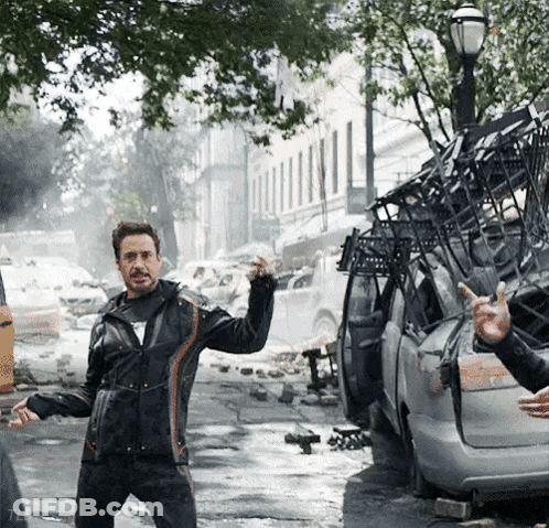 A Man In A Black Jacket Is Standing In Front Of A Destroyed Car And Giving The Middle Finger GIF