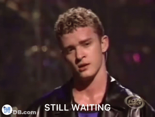 A Man In A Black Jacket Is Standing In Front Of A Sign That Says " Still Waiting " GIF