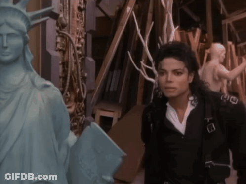 A Man In A Black Jacket Is Standing Next To A Statue Of The Liberty GIF