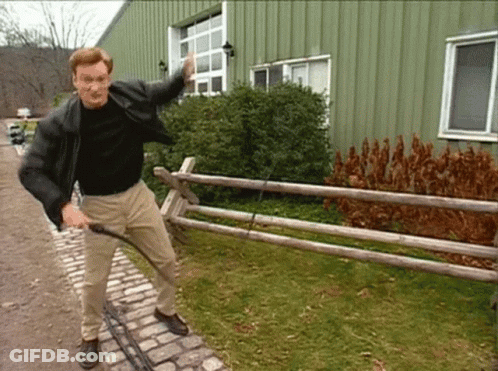 A Man In A Black Jacket Is Standing Next To A Wooden Fence Holding A Hose GIF