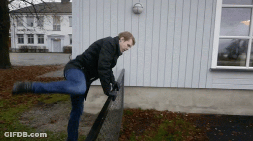 A Man In A Black Jacket Is Standing On One Leg On A Chain Link Fence GIF
