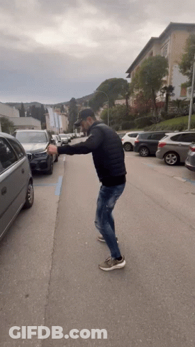 A Man In A Black Jacket Is Standing On The Side Of A Road GIF