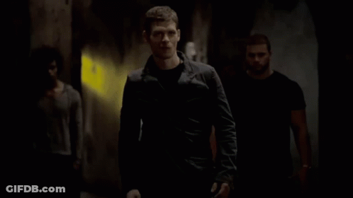 A Man In A Black Jacket Is Walking Down A Dark Hallway With Two Other Men . GIF