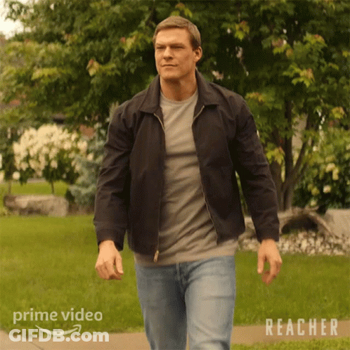 A Man In A Black Jacket Is Walking Down A Sidewalk In Front Of A Lush Green Field . GIF