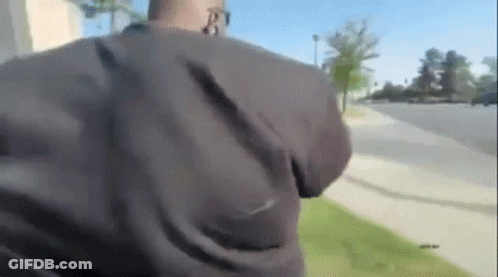 A Man In A Black Jacket Is Walking Down A Sidewalk While Talking On A Cell Phone . GIF