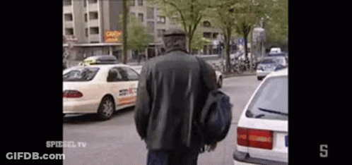 A Man In A Black Jacket Is Walking Down A Street Next To A Taxi . GIF