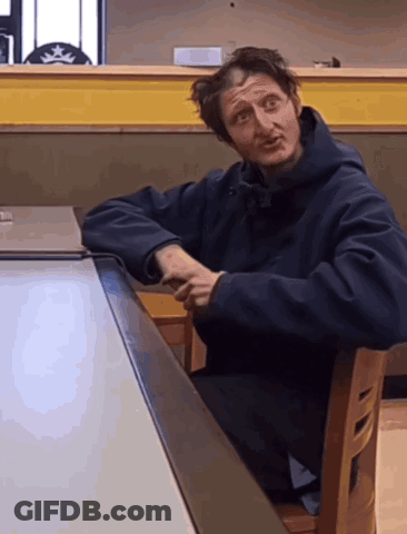 A Man In A Black Jacket Sits At A Table With His Arms Crossed GIF