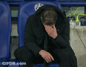 A Man In A Black Jacket Sits In A Blue Chair With His Head In His Hands GIF
