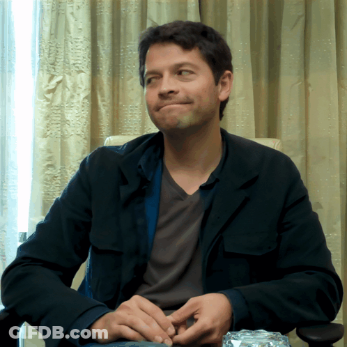 A Man In A Black Jacket Sits In A Chair With His Hands Folded GIF