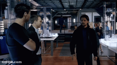 A Man In A Black Jacket Stands In A Room With Other Men And A Woman GIF
