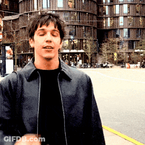A Man In A Black Jacket Stands In Front Of A Building That Says ' A ' On The Front GIF