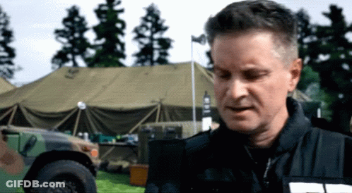 A Man In A Black Jacket Stands In Front Of A Military Vehicle With The Word Army On It GIF