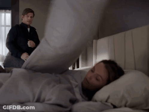 A Man In A Black Jacket Stands Next To A Man Laying In Bed GIF