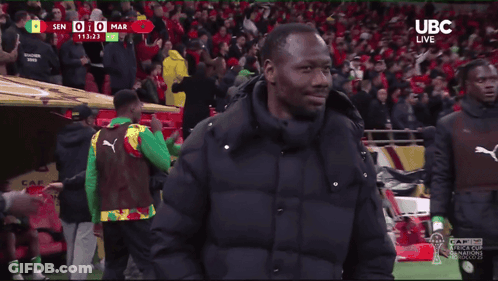 A Man In A Black Jacket Stands On A Soccer Field In Front Of A Crowd With Ubc Live Written On The Bottom GIF
