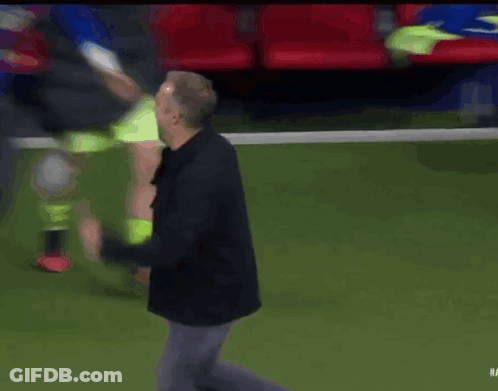 A Man In A Black Jacket Stands On A Soccer Field Surrounded By Players GIF