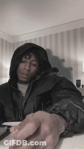 A Man In A Black Jacket With A Hood Is Sitting At A Table GIF