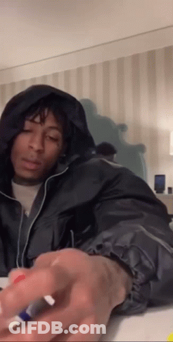 A Man In A Black Jacket With A Hood Is Sitting On A Bed Holding A Marker . GIF
