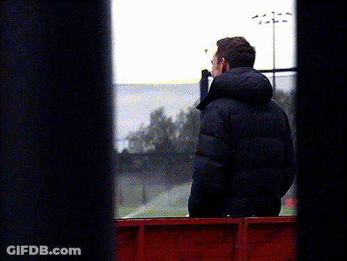 A Man In A Black Jacket With A Hood Looks Out A Window GIF