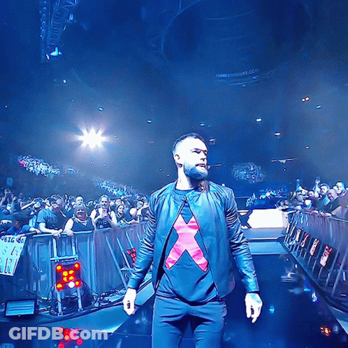 A Man In A Black Jacket With A Red X On His Shirt Stands On A Stage GIF