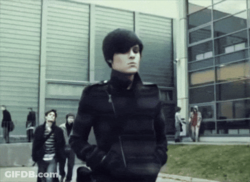A Man In A Black Jacket With His Hands In His Pockets Stands In Front Of A Building GIF