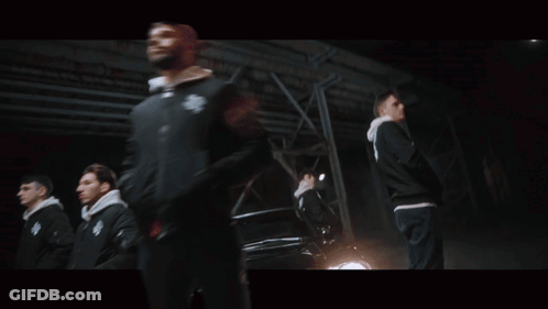 A Man In A Black Jacket With The Letter S On It Stands In Front Of A Car GIF