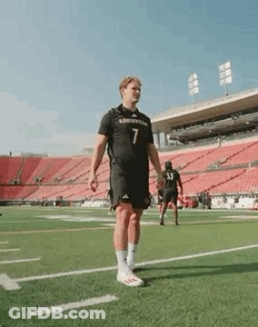 A Man In A Black Jersey Is Standing On A Football Field . GIF