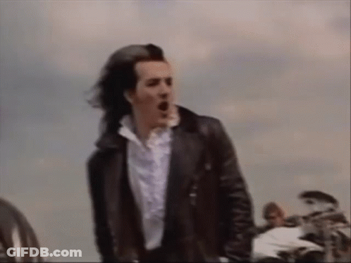 A Man In A Black Leather Jacket And White Shirt Is Standing In Front Of A Motorcycle . GIF