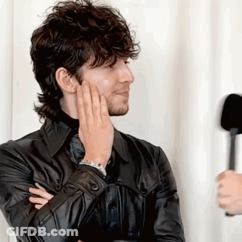 A Man In A Black Leather Jacket Is Holding His Hand To His Face While Talking Into A Microphone . GIF