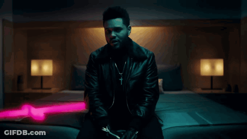 A Man In A Black Leather Jacket Is Sitting On A Bed With A Pink Light Behind Him GIF