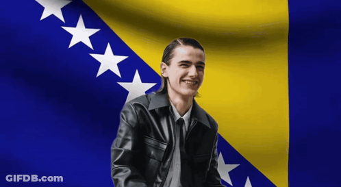 A Man In A Black Leather Jacket Is Smiling In Front Of A Flag GIF