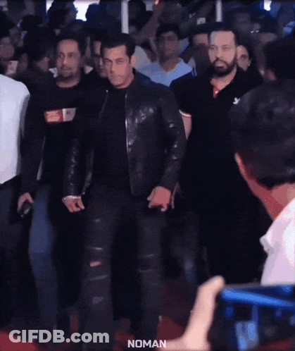 A Man In A Black Leather Jacket Is Walking On A Red Carpet With The Word Noman On The Bottom Right GIF