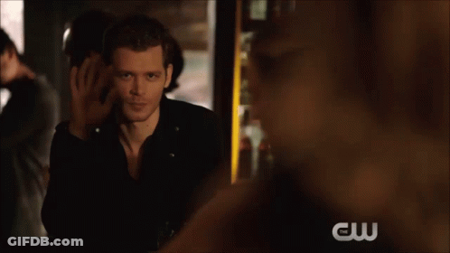 A Man In A Black Leather Jacket Sits With His Feet Up And A Cw Logo In The Background GIF
