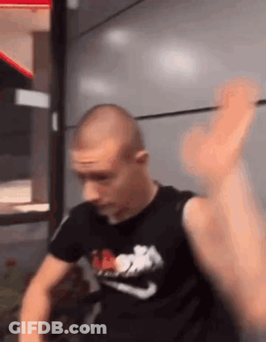 A Man In A Black Nike Shirt Is Standing In Front Of A Wall With His Hand Up . GIF
