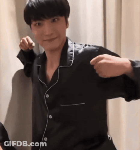 A Man In A Black Pajama Shirt Is Standing In Front Of A White Curtain And Making A Fist . GIF