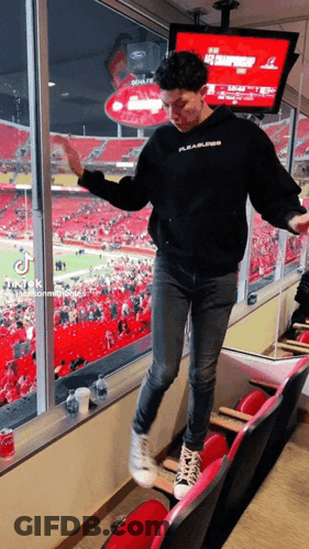 A Man In A Black Pleasures Hoodie Stands In A Stadium Watching A Football Game GIF