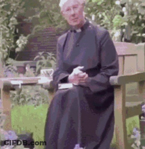 A Man In A Black Robe Is Sitting On A Bench In A Garden GIF