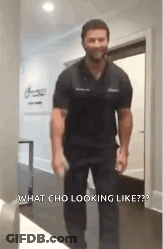 A Man In A Black Shirt And Black Pants Is Walking Down A Hallway With A Bottle In His Hand . GIF