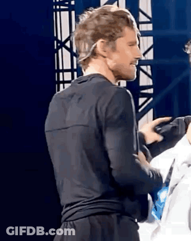 A Man In A Black Shirt And Black Shorts Is Standing On A Stage With A Microphone On His Head . GIF