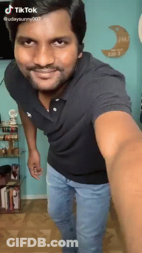A Man In A Black Shirt And Blue Jeans Is Taking A Selfie In A Room . GIF