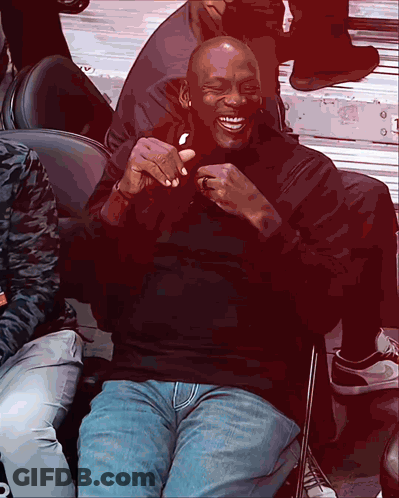 A Man In A Black Shirt And Blue Jeans Sits In A Chair And Laughs GIF