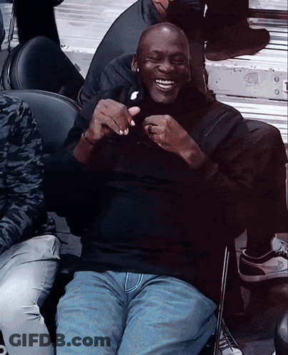 A Man In A Black Shirt And Blue Jeans Sits In A Chair And Smiles GIF