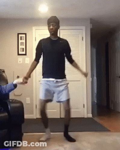 A Man In A Black Shirt And Blue Shorts Is Dancing In A Living Room . GIF