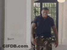 A Man In A Black Shirt And Camo Pants Is Walking In A Room . GIF