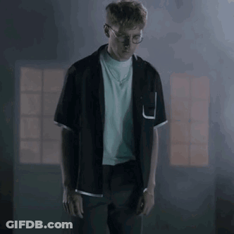 A Man In A Black Shirt And Glasses Is Standing In A Dark Room . GIF
