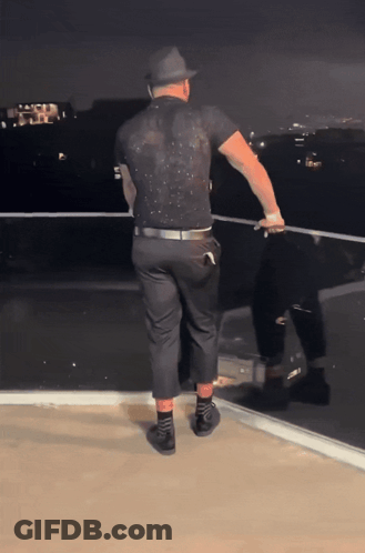 A Man In A Black Shirt And Hat Is Standing On A Ledge GIF