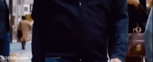 A Man In A Black Shirt And Jeans Is Standing In A Crowded Room . GIF